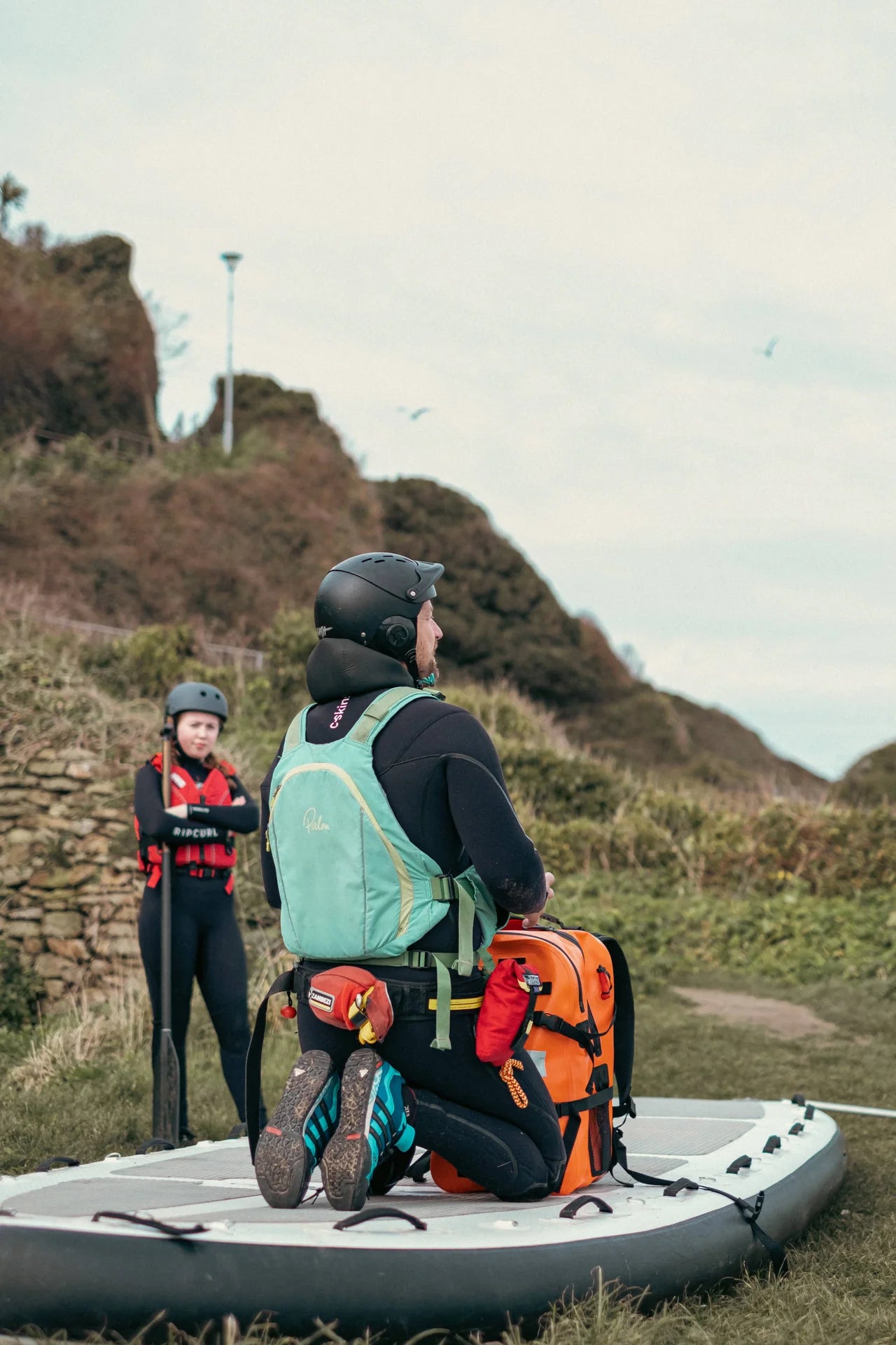 An instructor on a paddle board, talking to his students, he has an orange ULU bag